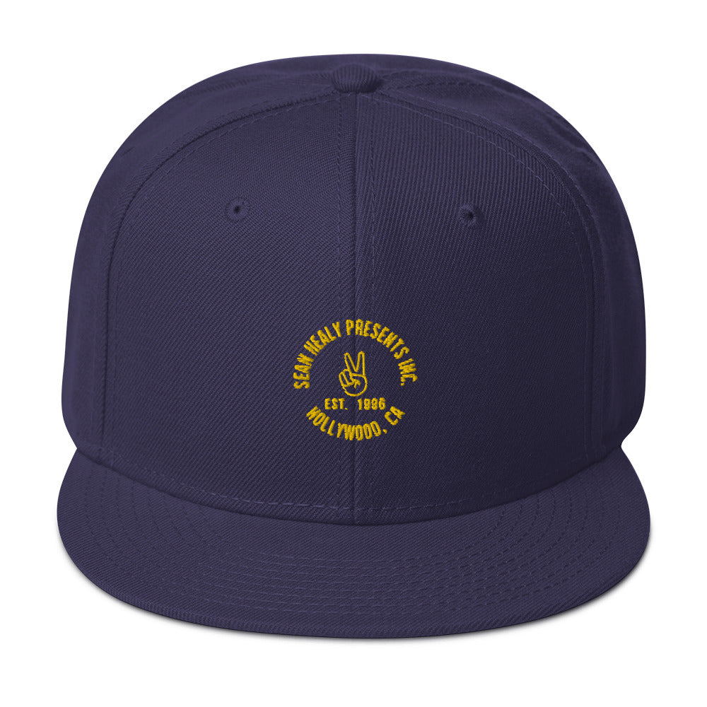 A navy blue snapback hat with a flat visor and a subtle grey under visor, featuring an embroidered peace sign and the text 'SHP PRESENTS EST. 2000 L.